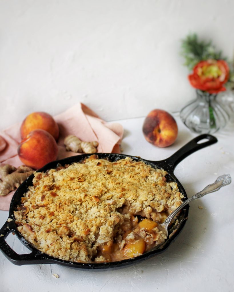 Ginger peach crisp in a cast iron skillet surrounded by peaches and flowers