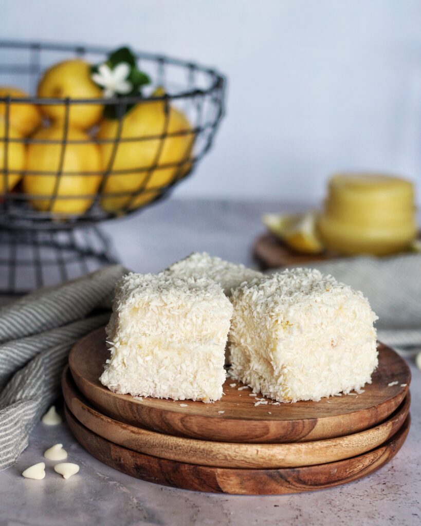 Three lemon white chocolate lamingtons on a wooden plate, lemons in a basket. 