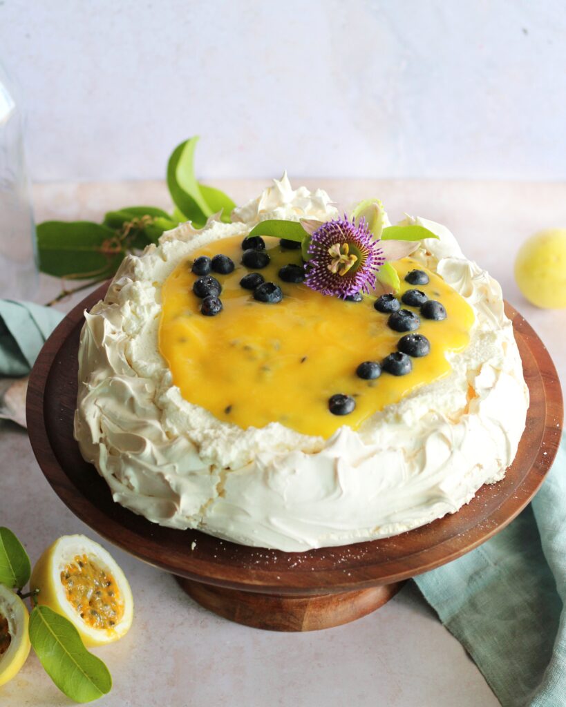 Pavlova on a cake stand, topped with passion fruit curd, fruit, and flowers 