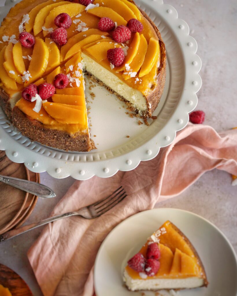 photo of mango coconut cheesecake, sliced, topped with raspberries and coconut. 